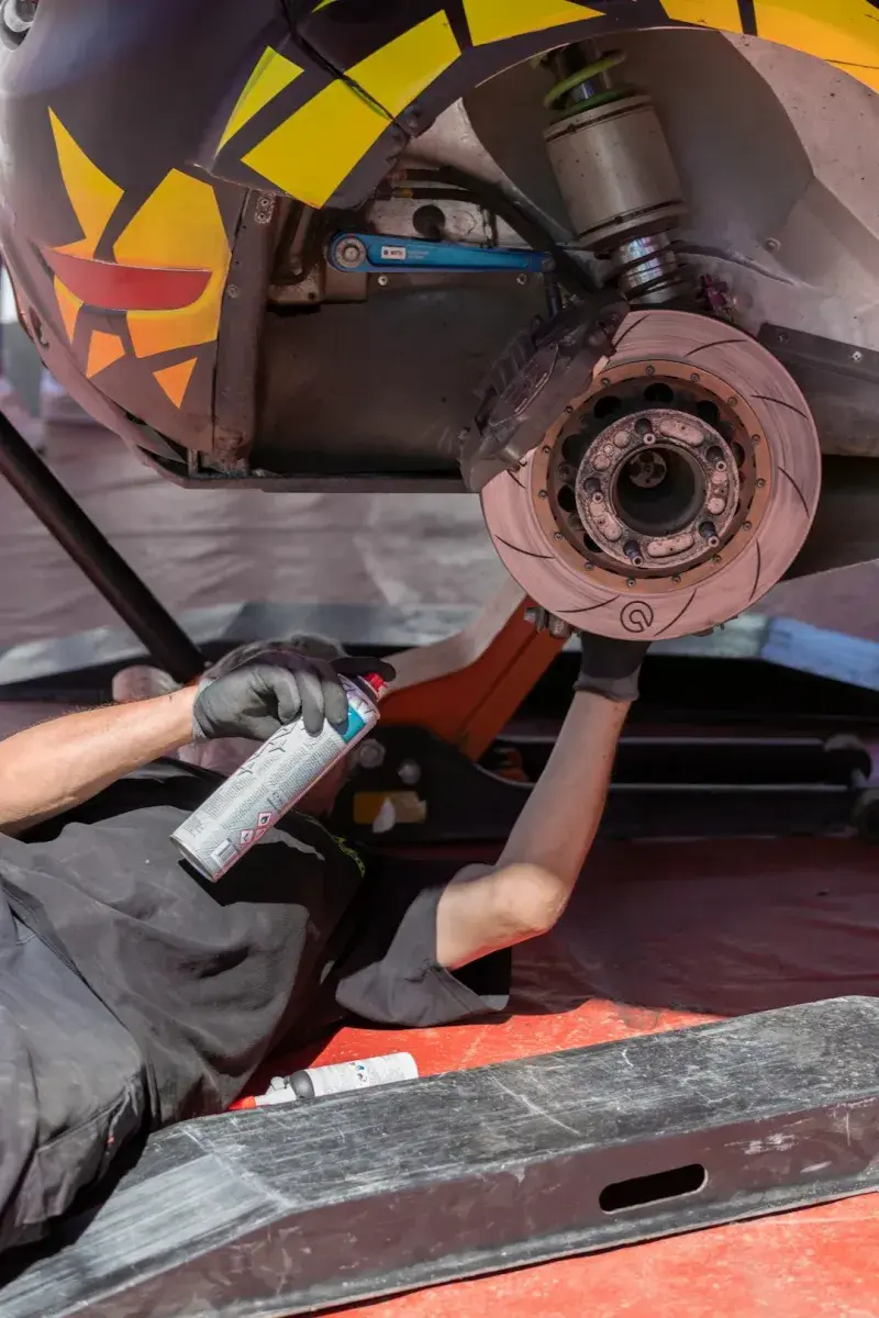 Technician spraying brake cleaner onto a slotted brake disc from under a raised car